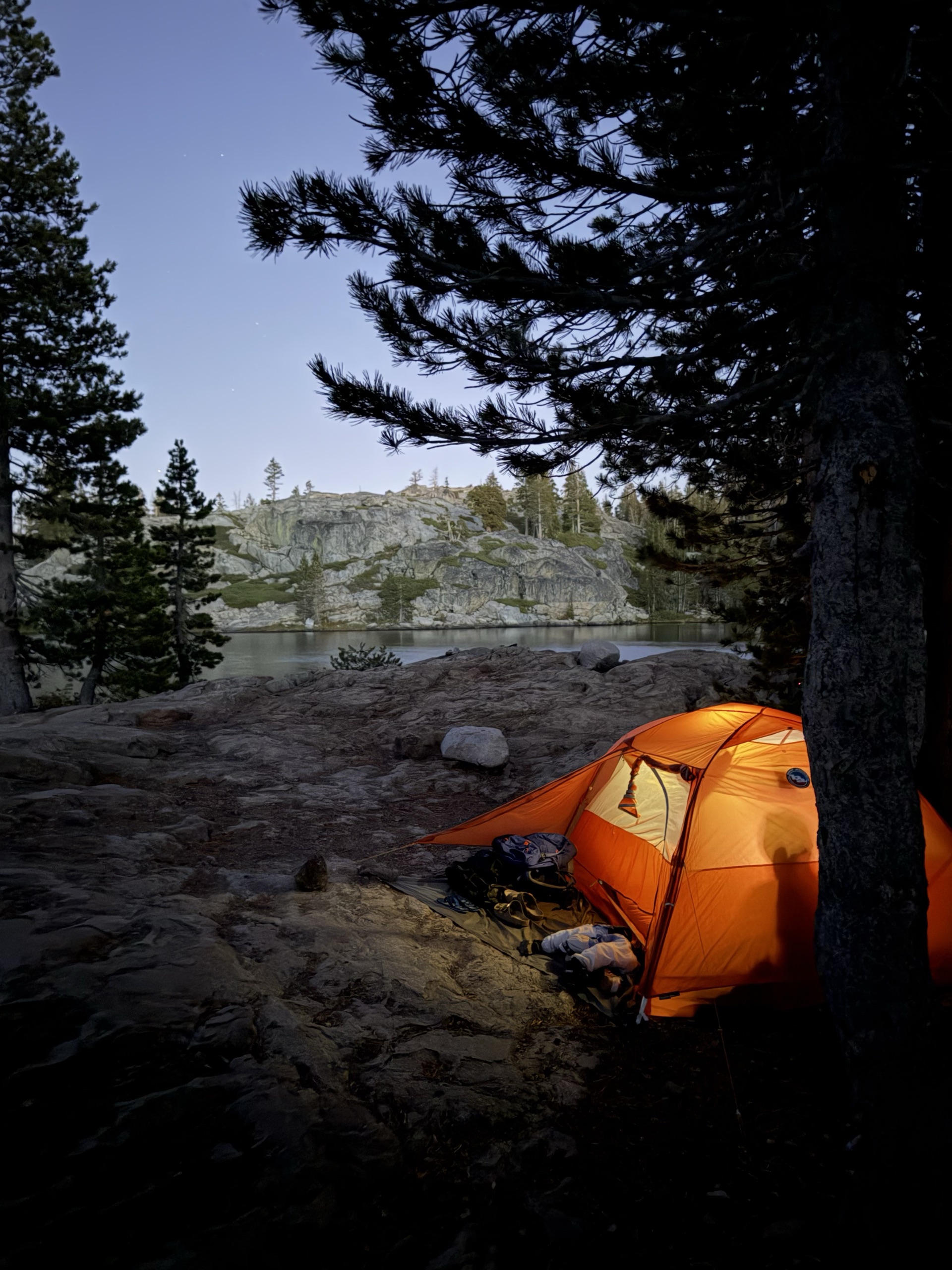 An orange tent glowing at dusk beside a granite-ringed lake