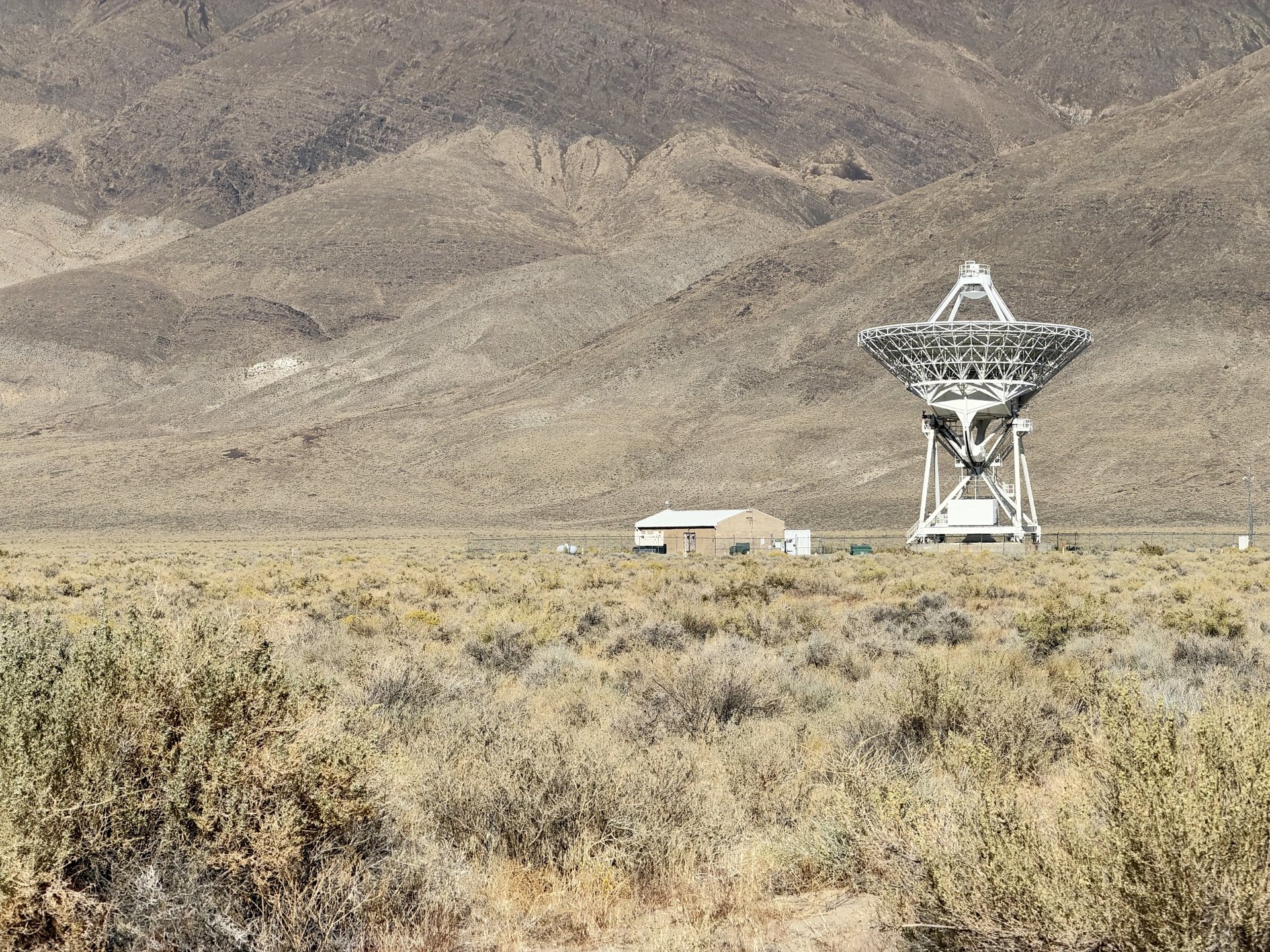 A white radio telescope dish and a small building below a bare desert mountainside
