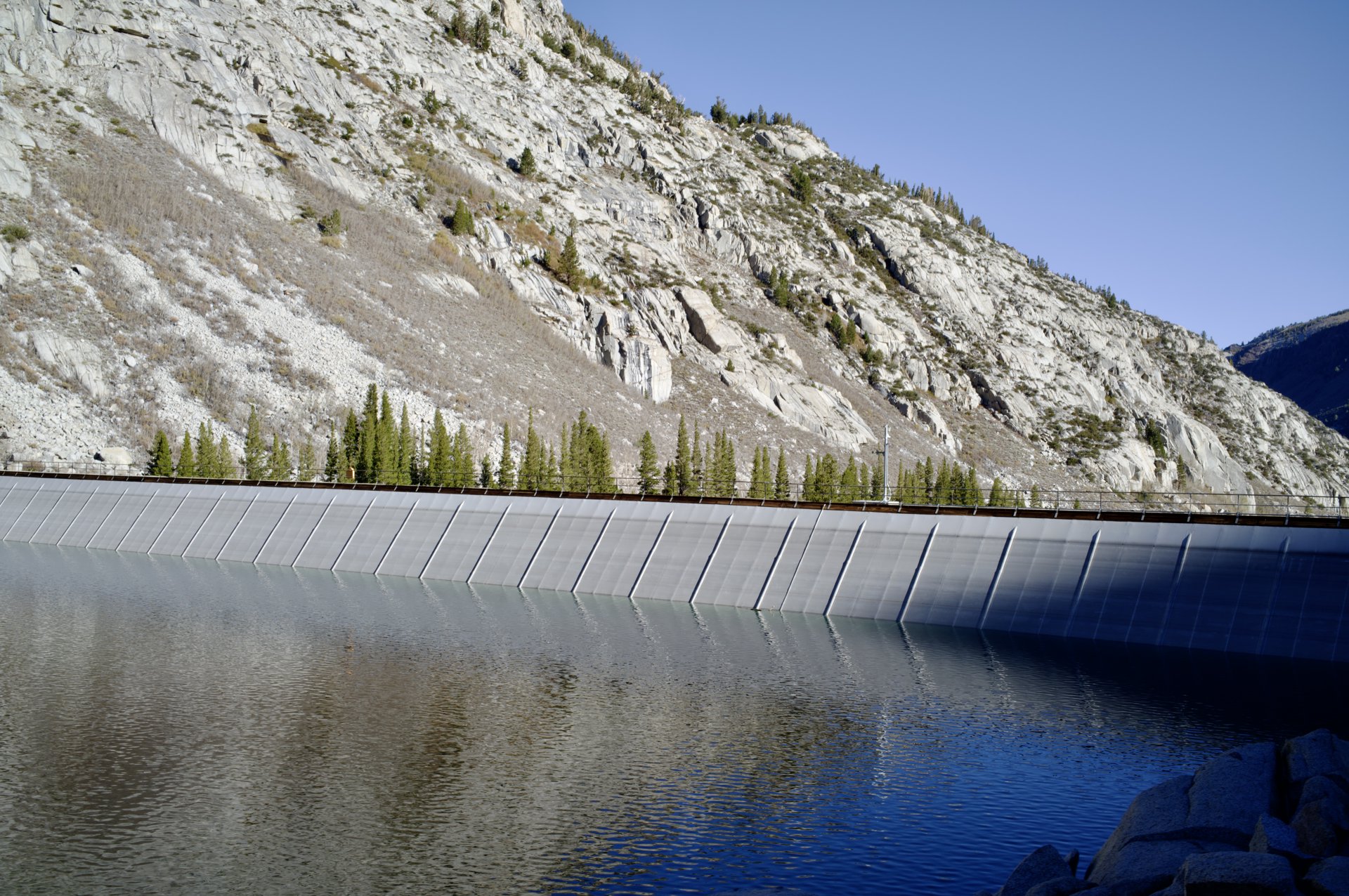 A curved concrete dam below a granite slope, reflected in the still water of a reservoir